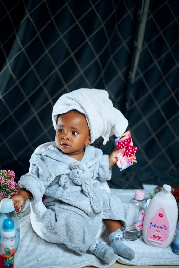 Cute baby in a bathrobe surrounded by skincare products, indoors.