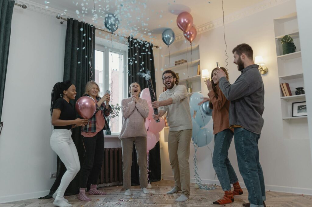 Excited friends celebrate a gender reveal with balloons and confetti indoors.