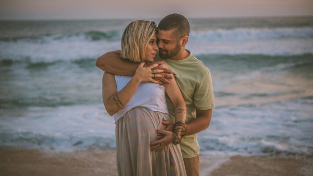 A couple embracing on the beach, celebrating pregnancy at sunset.