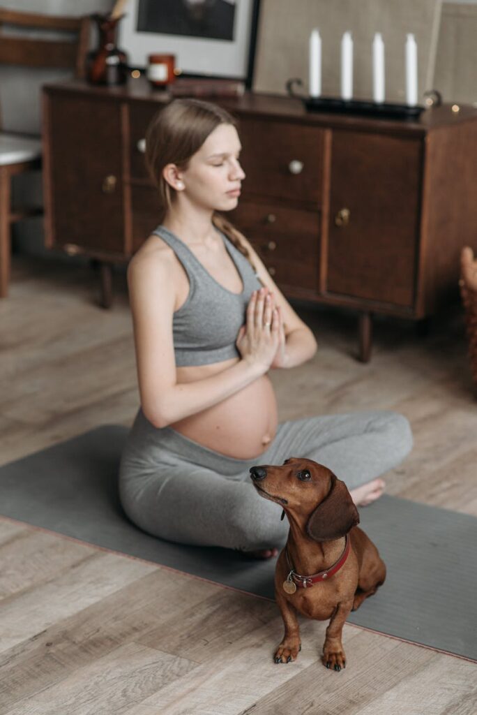 Pregnant woman in activewear meditating on yoga mat with a dachshund dog beside her indoors.