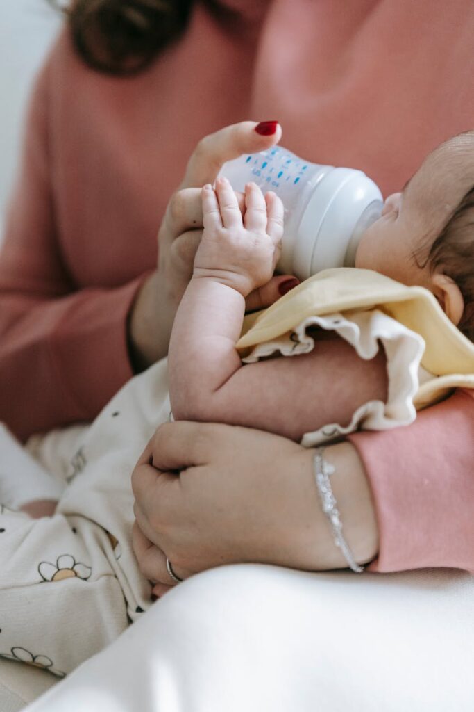 Crop unrecognizable mother in casual wear feeding crop adorable infant with bottle in arms while sitting in light room at home