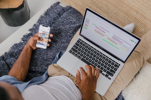 Man multitasking with phone and laptop on sofa, showcasing modern remote work lifestyle.
