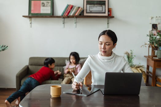 Mother multitasking with laptop and stylus alongside her children playing at home.