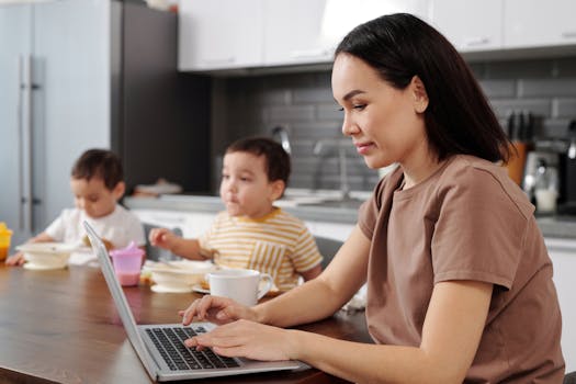Mother multitasking with laptop as children have breakfast on wooden table.