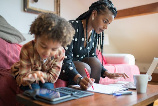A mother multitasking at home, working on a laptop while her child plays on a tablet nearby.